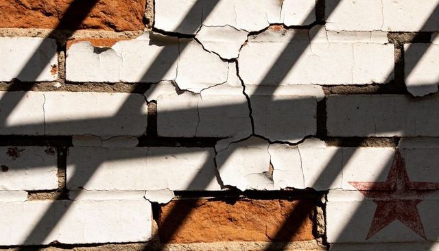 Weathered brick facade showing peeling white paint, exposed red bricks, diagonal shadows