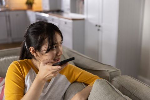 Asian woman speaking into smartphone while relaxing on sofa in contemporary open-plan living room