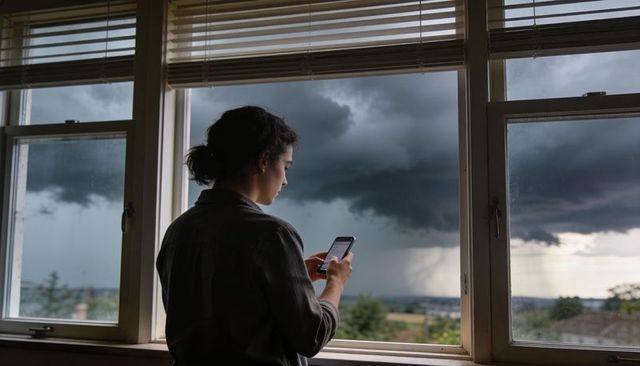 Woman watching distant thunderstorm through window while checking smartphone at home