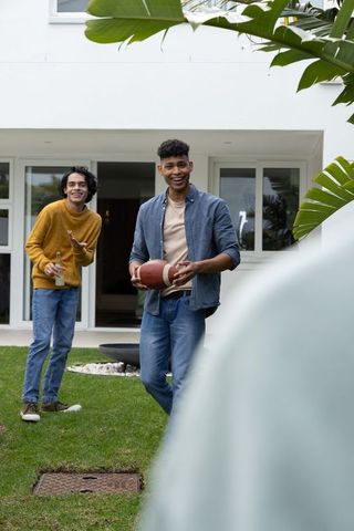 Smiling diverse friends playing catch with football on front lawn of modern home