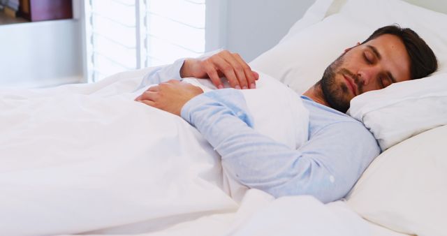 An image of a young man peacefully sleeping in a cozy white bed, captured in soft morning light. Perfect for use in articles or advertising related to sleep health, relaxation tips, or promoting bedding and home comfort products.