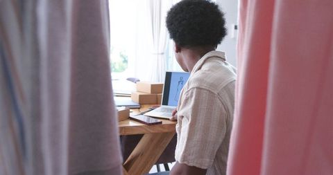 Designer reviewing laptop fashion sketch while packing parcels at minimal daylight workspace