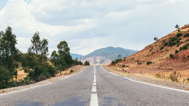 Long road stretching toward distant hills, ideal for themes around travel, adventure, freedom, and journey. Use for promoting road trips, motivational messages about the journey ahead, or as backdrops for automotive advertisements.
