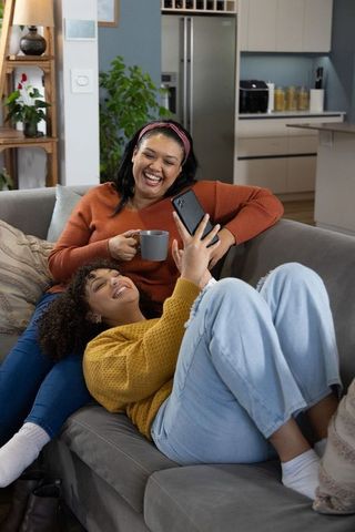 African american mother and teen daughter laughing and relaxing on sofa at home