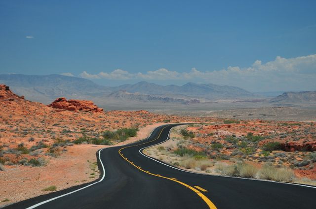 This image shows a winding road in a desert landscape leading towards distant mountains on a clear, sunny day. The road, flanked by red rocky terrain and sparse vegetation, curves gracefully, inviting travelers and adventurers. This image can be used for travel-related content, road trip promotion, outdoor adventure advertising, and motivational posters emphasizing wanderlust and exploration.