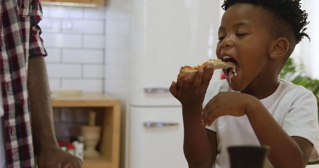 Child is enthusiastically eating a slice of pizza in a home kitchen. The scene captures a moment of joy during a mealtime, making it perfect for use in advertisements for household food products, family-oriented content, and childhood lifestyle visuals.