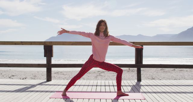 A woman is practicing yoga in the Warrior Pose on a beach boardwalk with the ocean and mountains in the background. This serene and peaceful setting is perfect for promotional materials for wellness programs, yoga retreats, outdoor fitness classes, or healthy lifestyle blogs and articles. It can also be used in advertisements for sports and activewear, meditation apps, or recreational activities related to nature and relaxation.