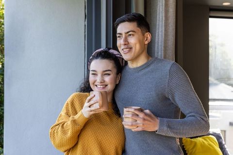 Young couple enjoying cozy morning coffee by window, holding ceramic mugs, smiling