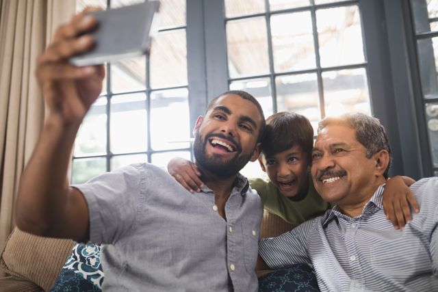 Multi-generation family smiling and taking a selfie together at home. Grandfather, father, and son are bonding and enjoying a happy moment in the living room. Ideal for use in family-oriented advertisements, social media posts, and articles about family relationships and technology.