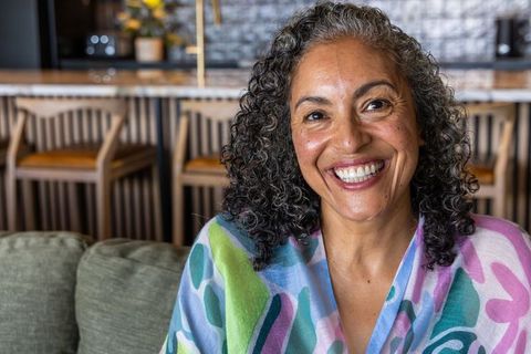 African american woman smiling on green sofa wearing colorful shawl, curly gray hair