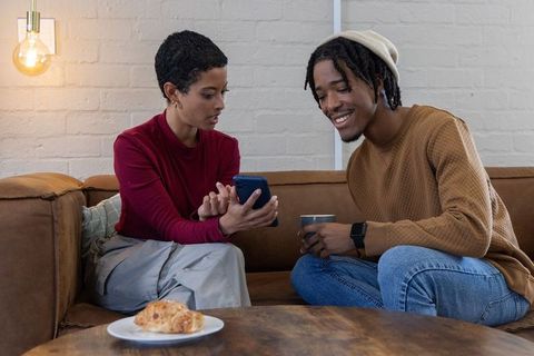 African american couple sharing smartphone and coffee on cozy living room couch