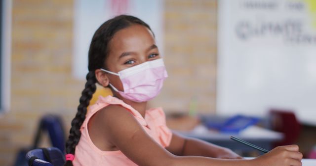 Schoolgirl with braided hair smiling while studying in classroom, sporting protective mask, representing safety and health protocols during pandemic. Ideal for depicting education, children's health, back-to-school themes, COVID-19 safety measures, and classroom environments.