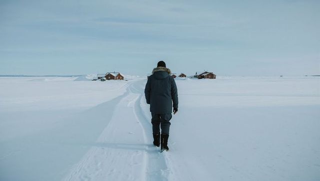Walking toward remote cabins across snowy tundra under overcast sky, solitary explorer