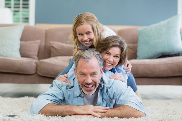 This image shows a joyful family of three lying on a carpet in their living room, with the parents and their daughter stacked on top of each other. They are all smiling, indicating a moment of bonding and togetherness. This image can be used for promoting family-oriented products, home decor, lifestyle blogs, or advertisements focusing on family values and happiness.