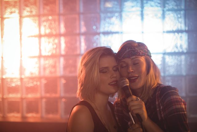 Two young women are singing into a microphone in a nightclub. They appear to be enjoying themselves, creating a lively and fun atmosphere. This image can be used for promoting nightlife events, music performances, or entertainment venues. It is also suitable for advertisements related to music, parties, and social gatherings.