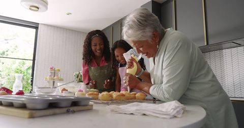 Multigenerational family decorating cupcakes with senior woman piping frosting in modern kitchen