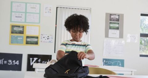Elementary student unpacking backpack in bright classroom with math posters and notebooks