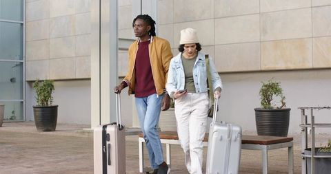 Young travelers walking with rolling suitcases and checking phone by modern plaza