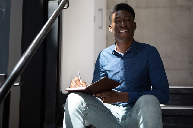 This image depicts a young African American businessman sitting on a staircase in a modern office environment, smiling while taking notes in a notebook. Ideal for use in business-related content, corporate training materials, articles on workplace diversity, and promotional materials for creative workspaces.