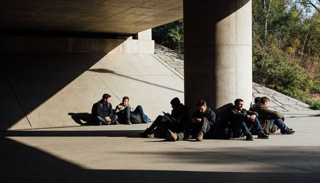 Young men resting under concrete overpass, leaning on pillar with backpacks and phones