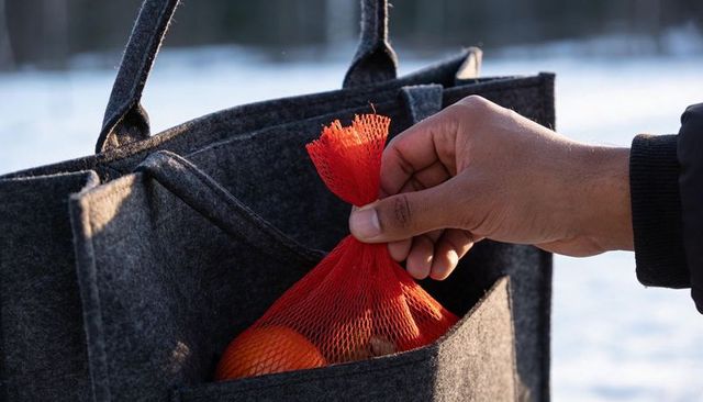 Hand placing red mesh bag of oranges into gray felt tote pocket in snowy winter outdoors