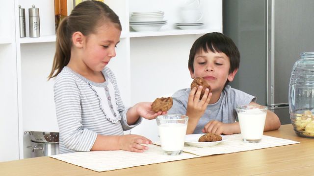 Young siblings are sitting at a kitchen table, eagerly eating cookies and drinking milk. They appear to be enjoying a sweet treat together, showcasing family bonding and happiness. Suitable for ads related to childhood, family time, home kitchens, snack food brands, and parent-focused products.