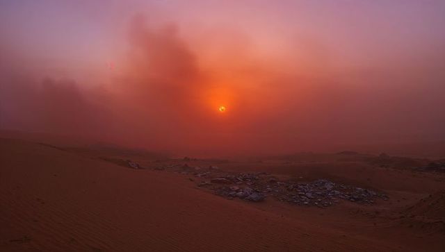 Glowing Sun Bathing Rippled Desert Dunes in Fiery Dust Haze with Scattered Rocks