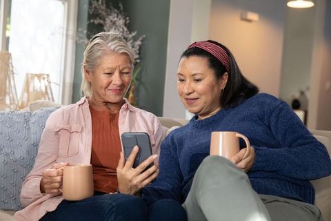 Two mature women sharing smartphone and coffee while relaxing together on sofa