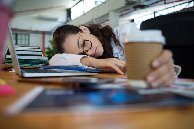Female graphic designer sleeping at her desk in an office, holding a disposable cup. Ideal for illustrating workplace fatigue, stress, overwork, and the need for breaks. Useful for articles on work-life balance, productivity, and mental health in the workplace.