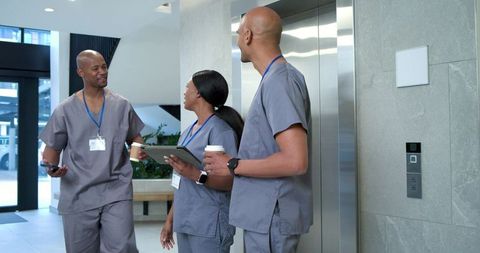 African american medical team chatting in hospital lobby by elevator holding tablet phone