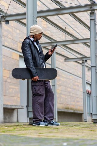 African american skater standing under glass canopy holding skateboard checking phone