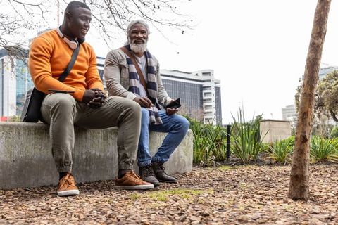 African american men sitting on bench chatting and using smartphone in urban park