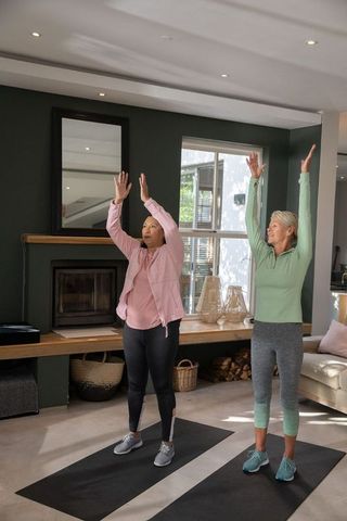 Two women practicing morning stretching routine on yoga mats in bright living room