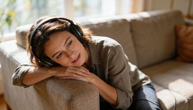 Woman relaxing on sofa wearing headphones in sunlit cozy living room, contemplating