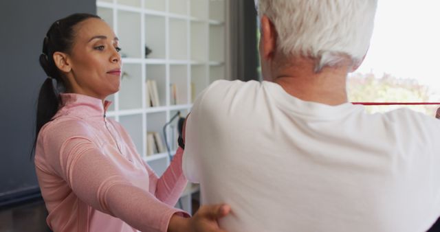 Female personal trainer providing guidance to a senior man using a resistance band for exercises. Perfect for illustrating themes related to fitness, personal training, senior health, physical therapy, rehabilitation, and active living in older adults. Useful for blogs, websites, and marketing materials promoting fitness services for seniors.