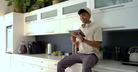 African american man sitting on kitchen counter using tablet and wearing headphones, smiling