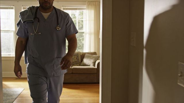 Home healthcare nurse walking into living room in blue scrubs with stethoscope, towel