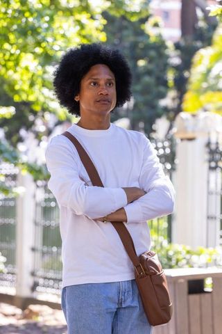 African american man standing with arms crossed in sunlit park wearing crossbody bag