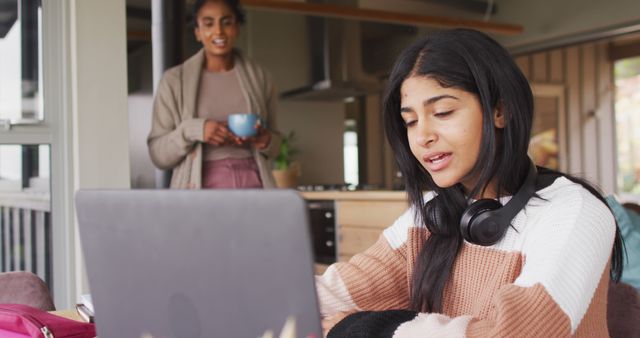Teen girl focused on studying with laptop, headphones around her neck. Background features mother holding coffee cup, observing. Ideal for themes related to home education, family support, academic activities, remote learning, and teenage lifestyle.