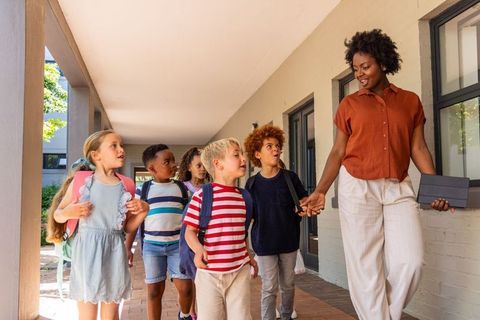 Female teacher leading diverse elementary students walking on sunny school corridor