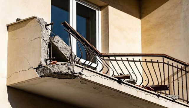 Damaged balcony exposing rusted rebar and bent railing on cracked stucco facade