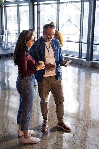 Networking colleagues viewing tablet in sunlit modern atrium with coffee cups and lanyards