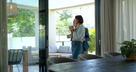 Diverse female couple enjoying morning coffee by sliding glass door in modern living room