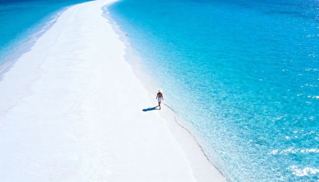 Solo woman walking along pristine sandbar with turquoise sea and white sunhat