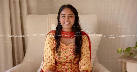 Smiling woman in yellow floral kurta with red dupatta seated in cream armchair home portrait