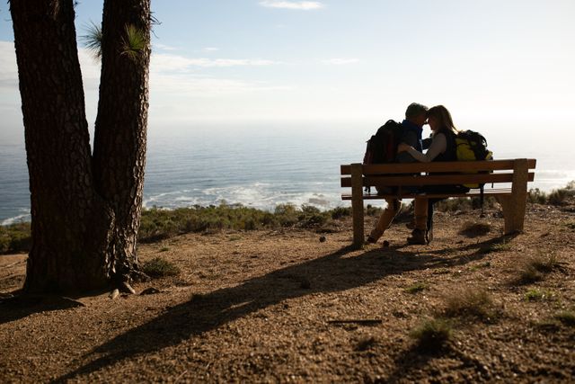 Side view of a happy senior Caucasian couple enjoying time in nature together, hiking in mountains, taking a break, sitting on a bench looking at each other, embracing with sea in the background.