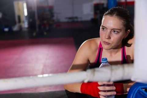 Female boxer leaning on ring ropes holding water bottle wearing red hand wraps taking break