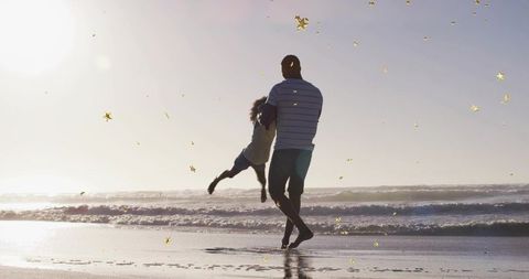 Man swinging child on sunlit beach at sunset with golden star confetti and silhouettes