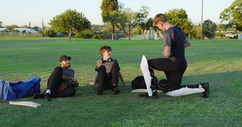 Sunset cricket teammates resting on grass while teen fastening white batting pads