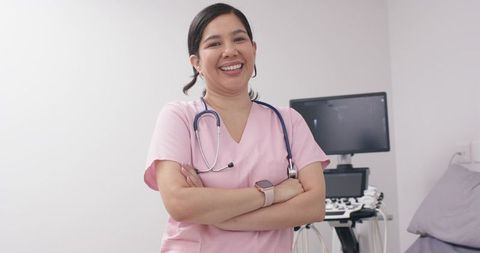 Smiling nurse wearing pink scrubs and stethoscope standing by ultrasound monitor in clinic